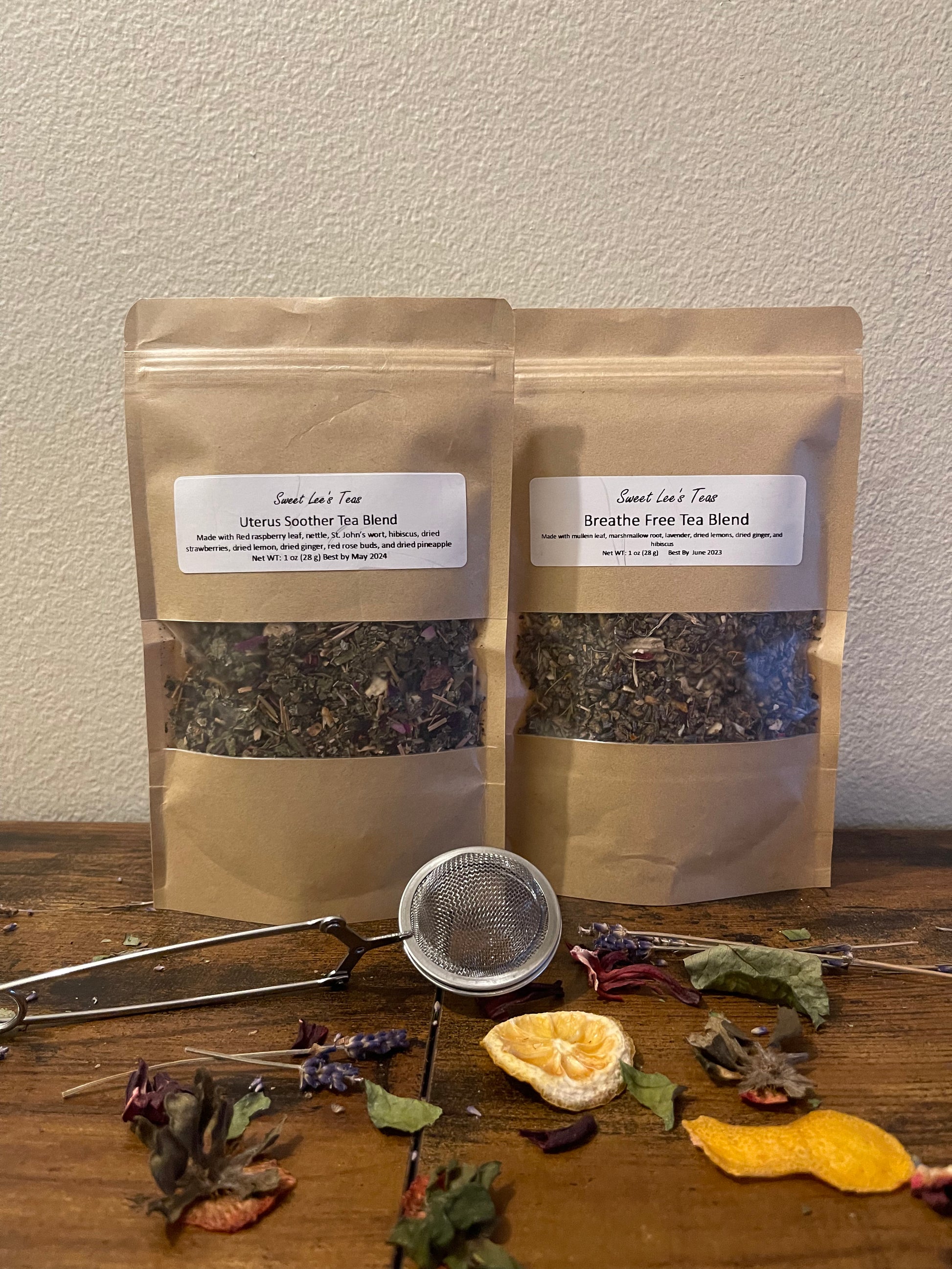 Two kraft bags of Sweet Lee’s Teas blends—Uterus Soother Tea Blend and Breathe Free Tea Blend—displayed on a wooden surface with a mesh tea infuser, dried citrus, and herbal ingredients, highlighting the natural and loose-leaf tea selection.