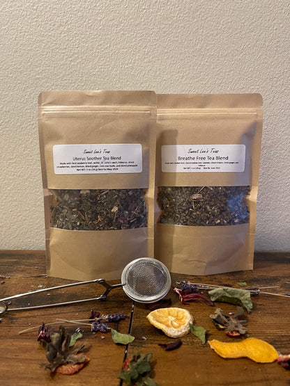 Two kraft bags of Sweet Lee’s Teas blends—Uterus Soother Tea Blend and Breathe Free Tea Blend—displayed on a wooden surface with a mesh tea infuser, dried citrus, and herbal ingredients, highlighting the natural and loose-leaf tea selection.