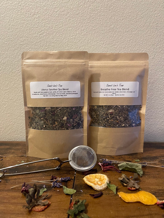 Two kraft bags of Sweet Lee’s Teas blends—Uterus Soother Tea Blend and Breathe Free Tea Blend—displayed on a wooden surface with a mesh tea infuser, dried citrus, and herbal ingredients, highlighting the natural and loose-leaf tea selection.
