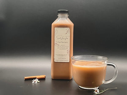 Sweet Lee’s Spiced Chai Latte in a clear bottle, displayed alongside a steaming glass cup of chai, a cinnamon stick, and a delicate white flower, set against a sleek black background.