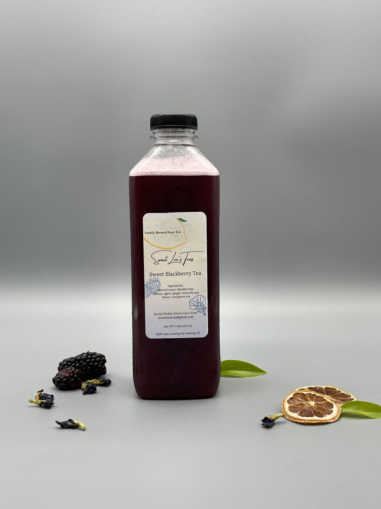 A bottle of Sweet Blackberry Tea from Sweet Lee’s Teas, surrounded by fresh blackberries, dried citrus slices, and butterfly pea flowers, highlighting its natural ingredients and deep berry flavor.