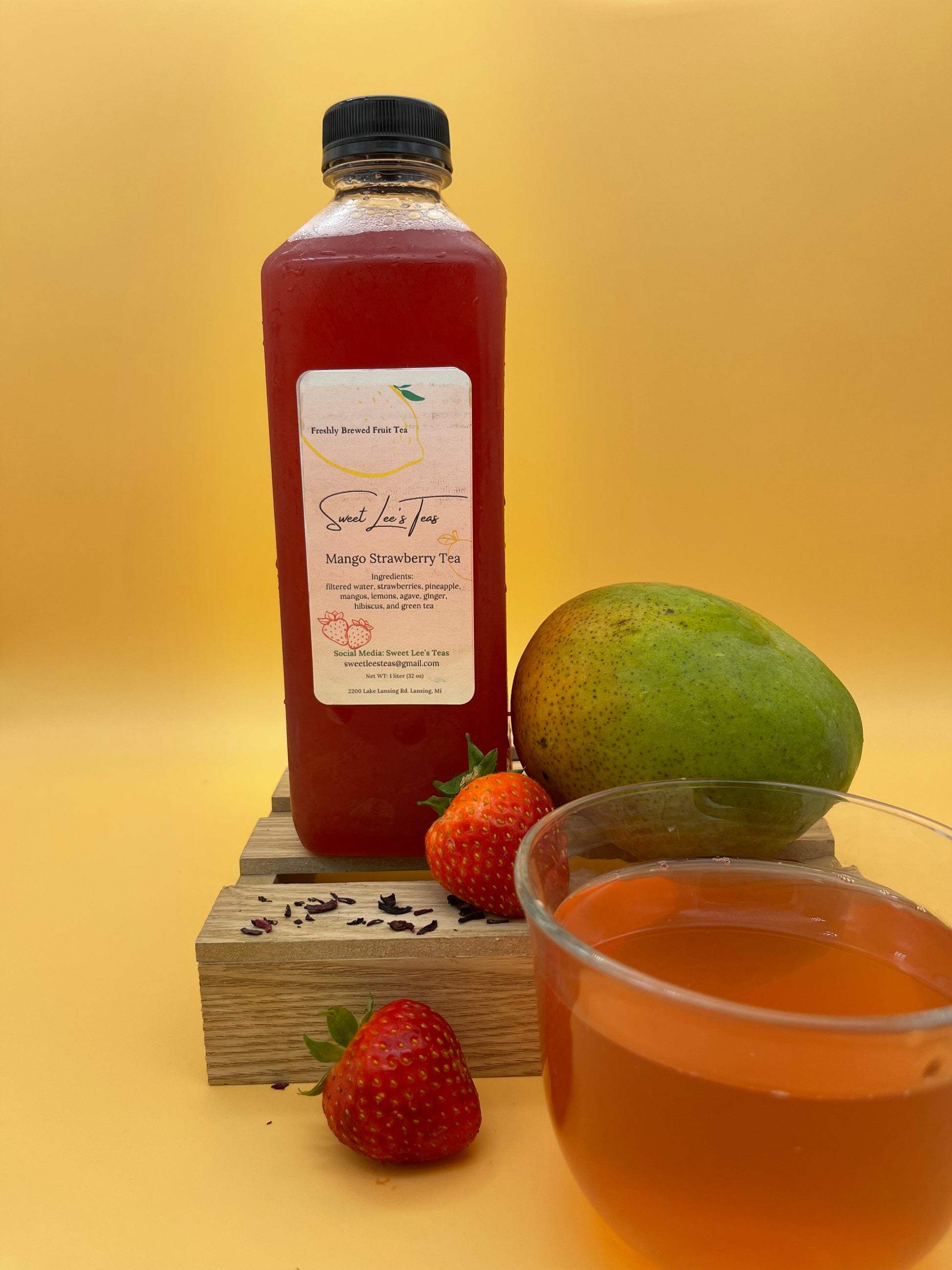 Sweet Lee’s Mango Strawberry Tea in a clear bottle, displayed on a wooden stand with fresh strawberries, a ripe mango, and a cup of brewed tea, set against a warm yellow background.