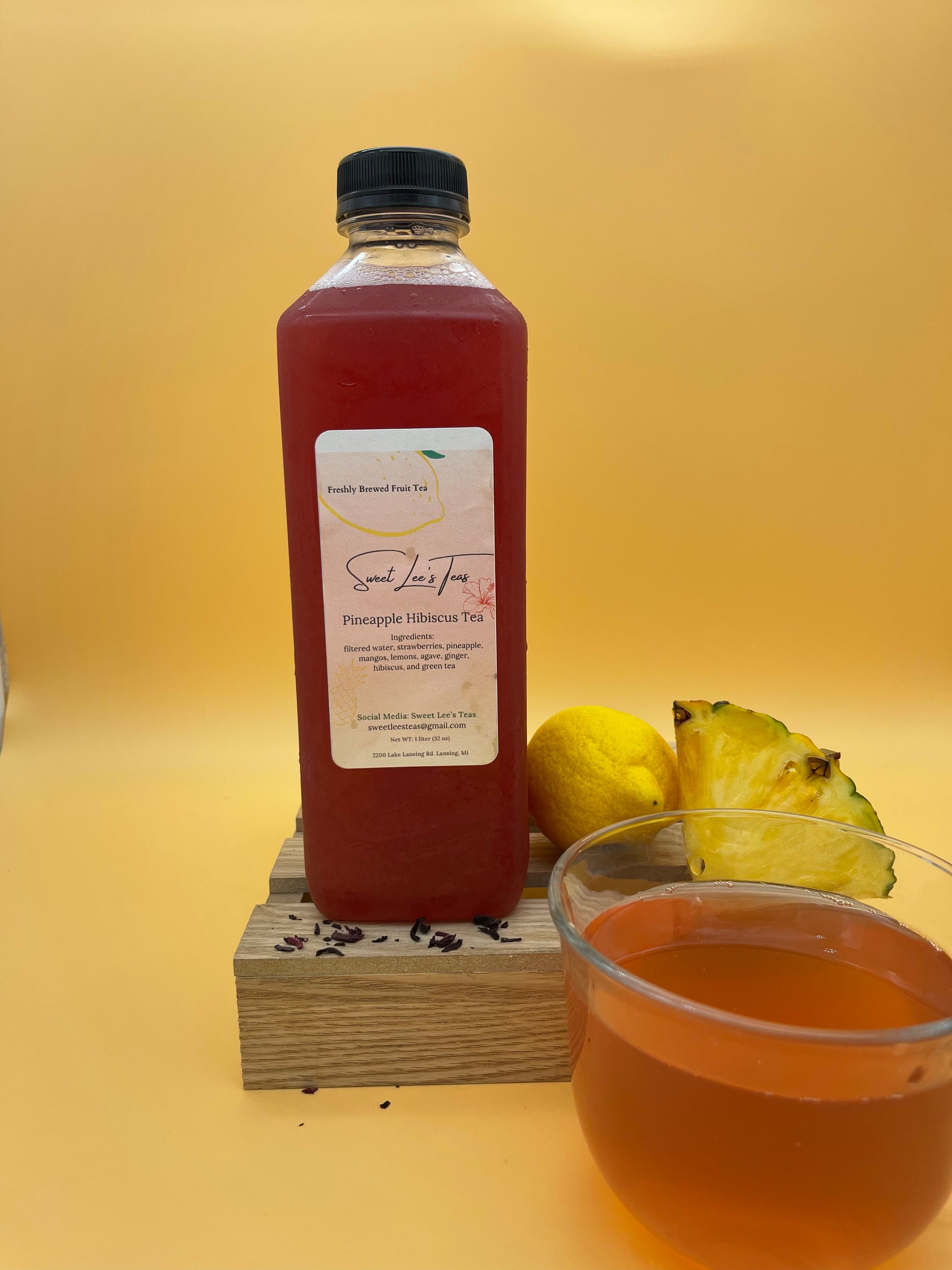 Sweet Lee’s Pineapple Hibiscus Tea in a clear bottle, displayed on a wooden stand with fresh pineapple, lemon, and a cup of brewed tea, set against a vibrant yellow background.