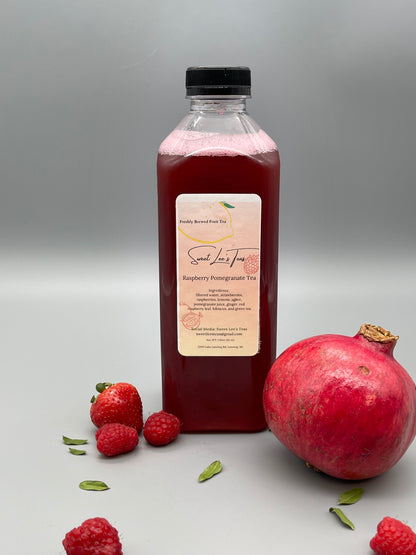Sweet Lee’s Raspberry Pomegranate Tea in a clear bottle, surrounded by fresh raspberries, strawberries, pomegranate, and green tea leaves, set against a neutral gray background.
