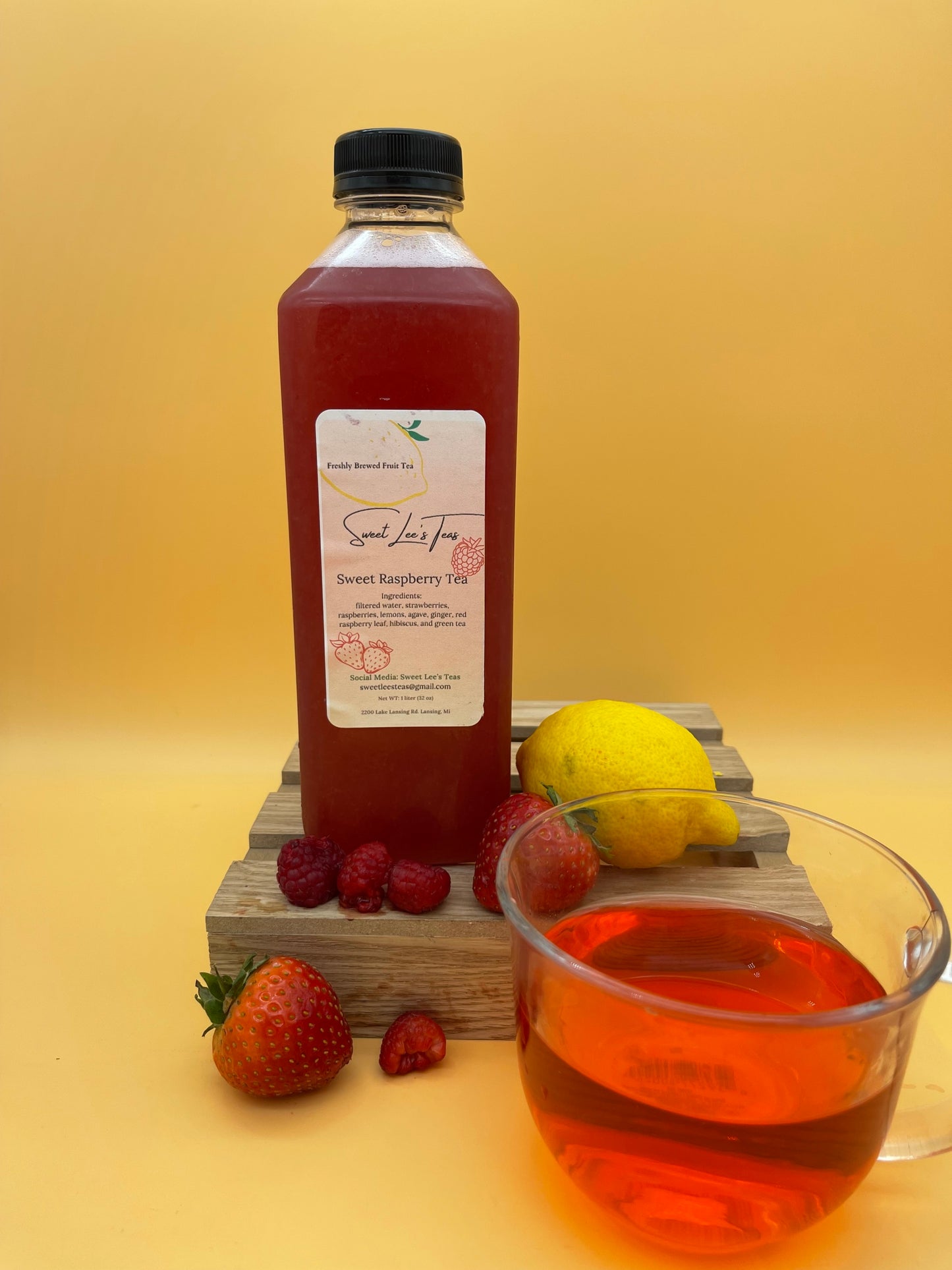 Sweet Lee’s Sweet Cherry Tea in a clear bottle, displayed with fresh raspberries, strawberries, a lemon, and a glass of vibrant red brewed tea on a wooden stand, set against a warm yellow background.