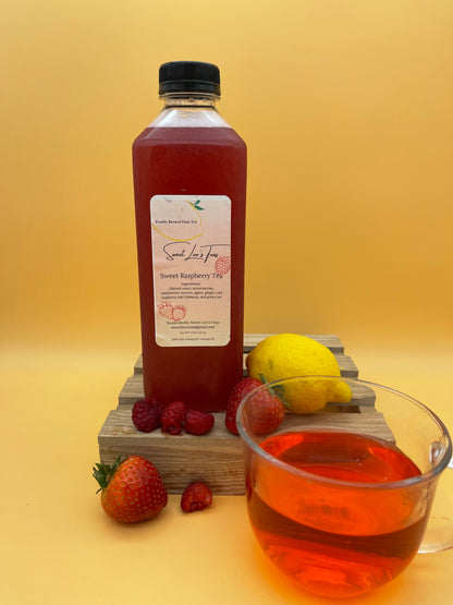 Sweet Lee’s Sweet Cherry Tea in a clear bottle, displayed with fresh raspberries, strawberries, a lemon, and a glass of vibrant red brewed tea on a wooden stand, set against a warm yellow background.
