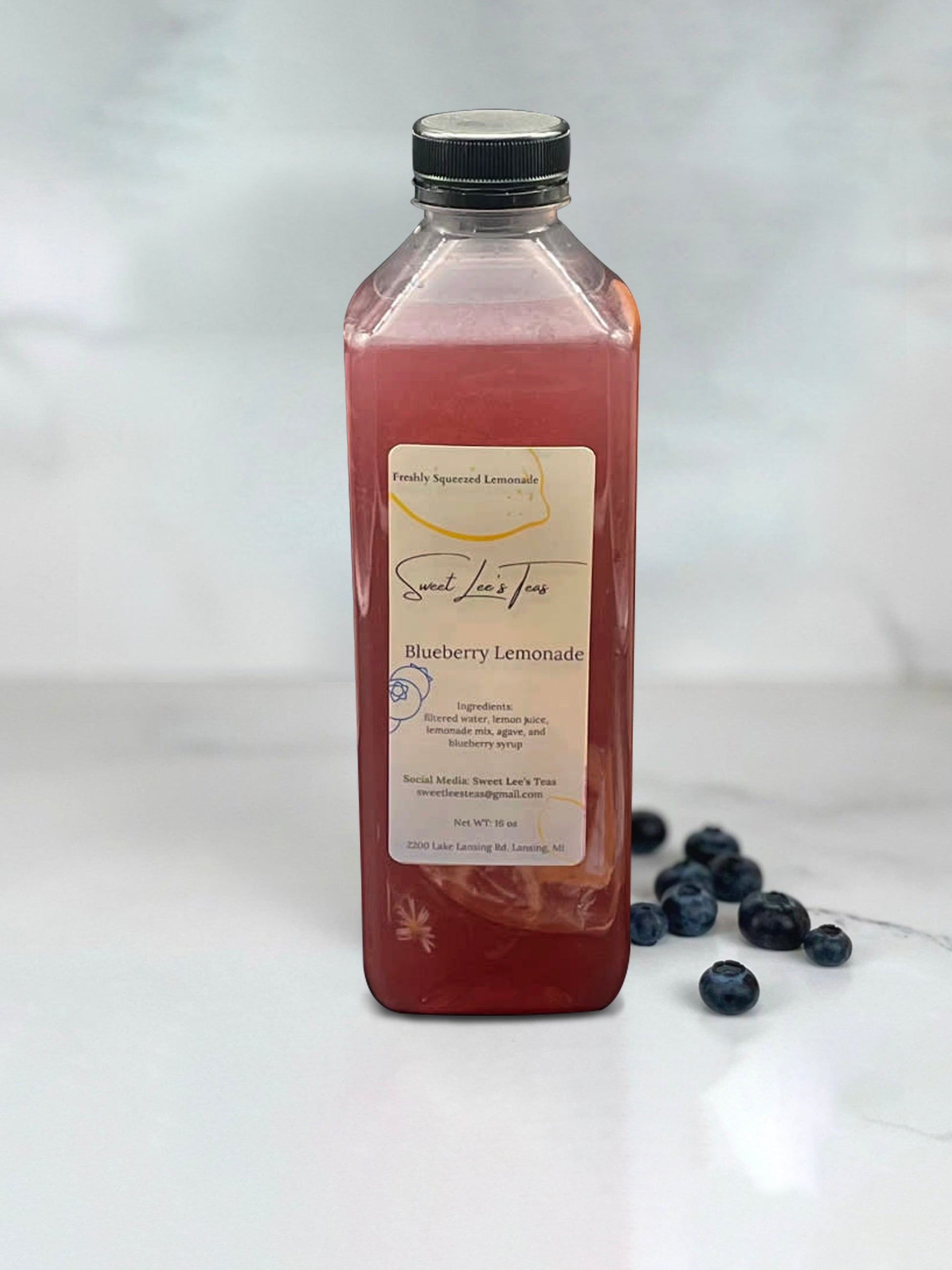 Sweet Lee’s Blueberry Lavender Lemonade in a clear bottle, placed on a marble surface with fresh blueberries scattered nearby, showcasing its vibrant color and refreshing citrus-berry blend.